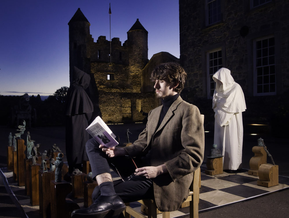 Adult man sitting on a chair reads from a book, behind two people in hoods stand on a large chessboard with wooden pieces, a castle and deep-blue sky is seen in the background.